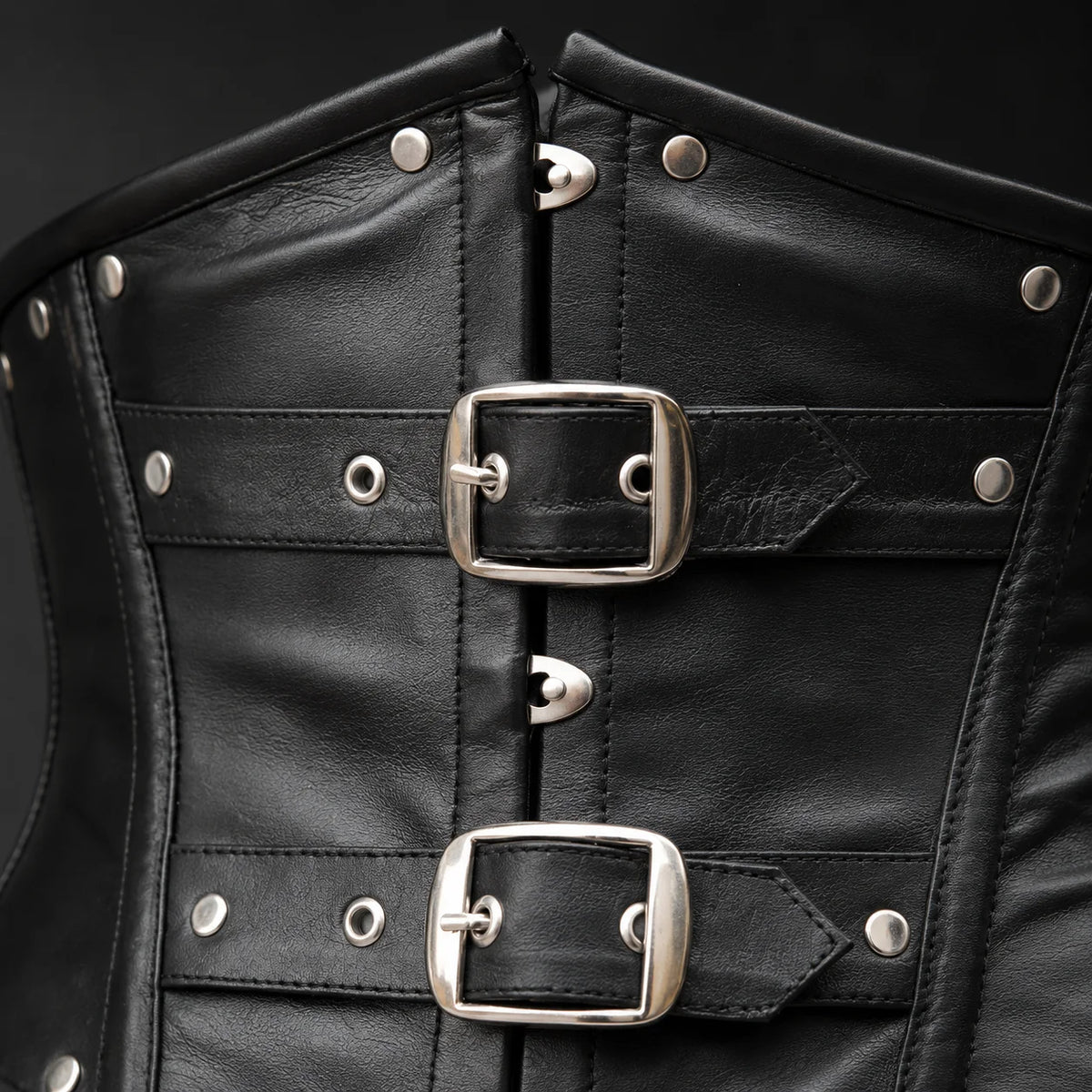 Close-up of black cowhide leather corset showing twin buckle hardware, studs, eyelets, stitching and leather texture.