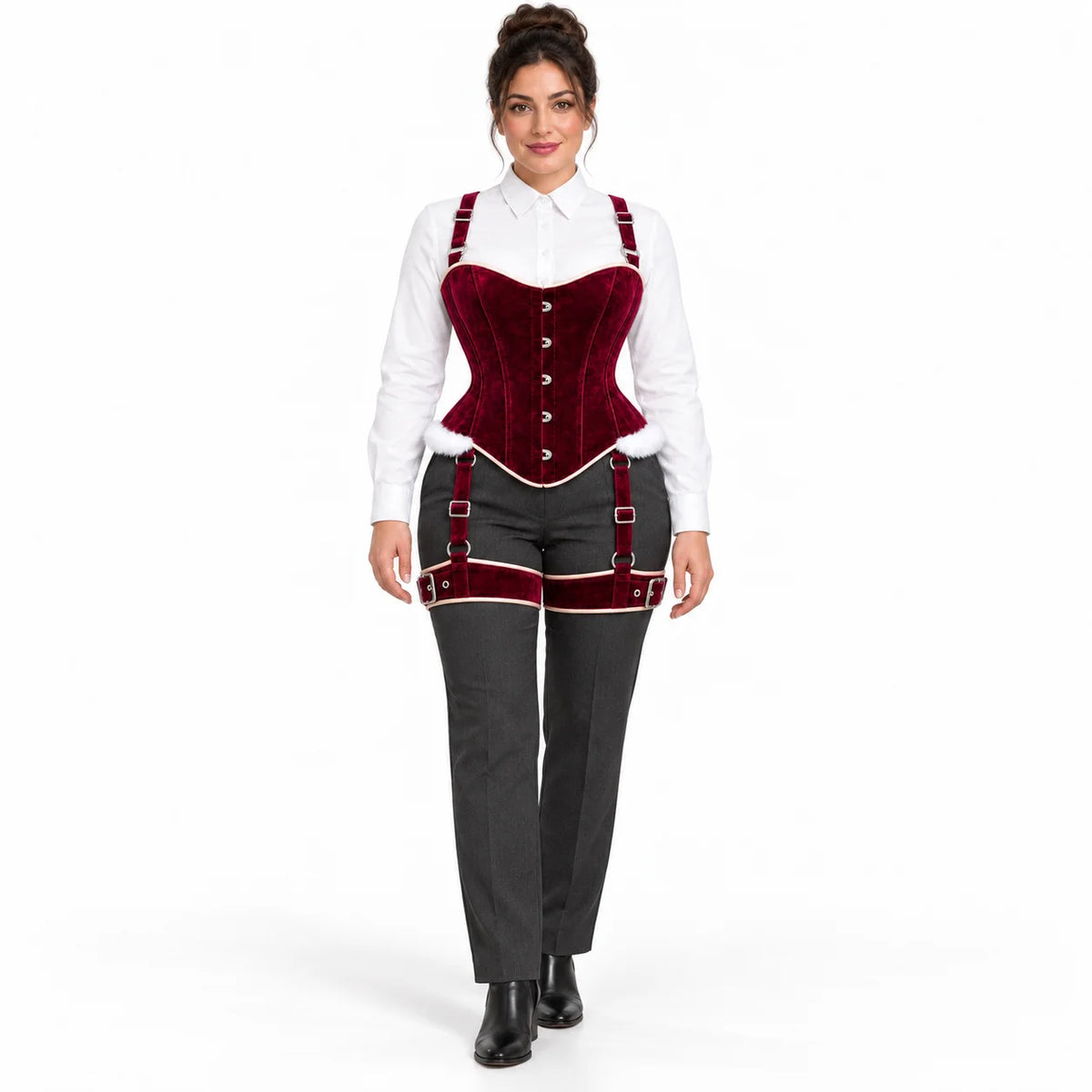 Full-length studio view of woman styling a burgundy velvet corset and matching thigh harness over grey trousers.