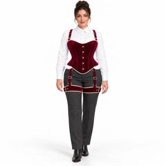 Full-length studio view of woman styling a burgundy velvet corset and matching thigh harness over grey trousers.