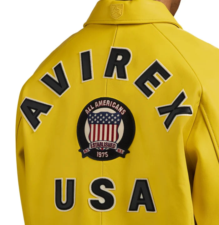 Close-up rear view of Avirex yellow bomber showing black AVIREX lettering, heritage crest and USA appliqué