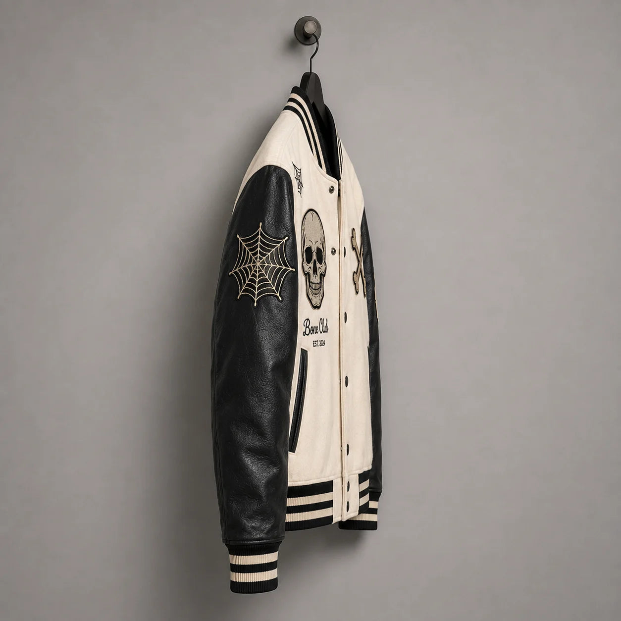 Side hanger view of Bone Club varsity jacket highlighting black faux leather sleeve and embroidered spider-web patch