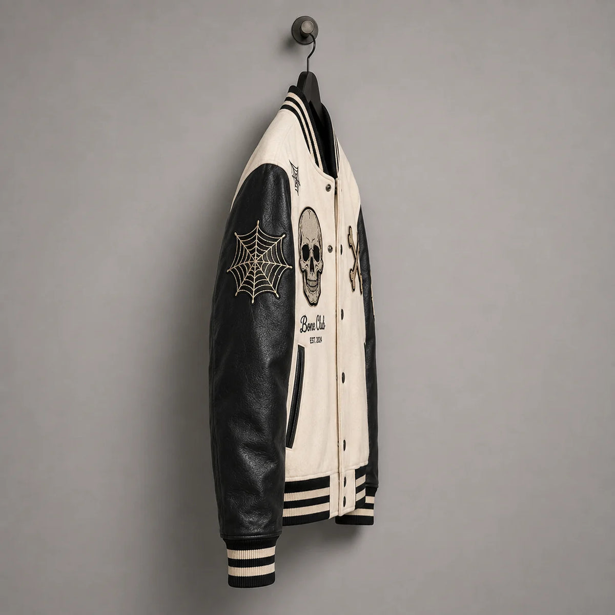 Side hanger view of Bone Club varsity jacket highlighting black faux leather sleeve and embroidered spider-web patch
