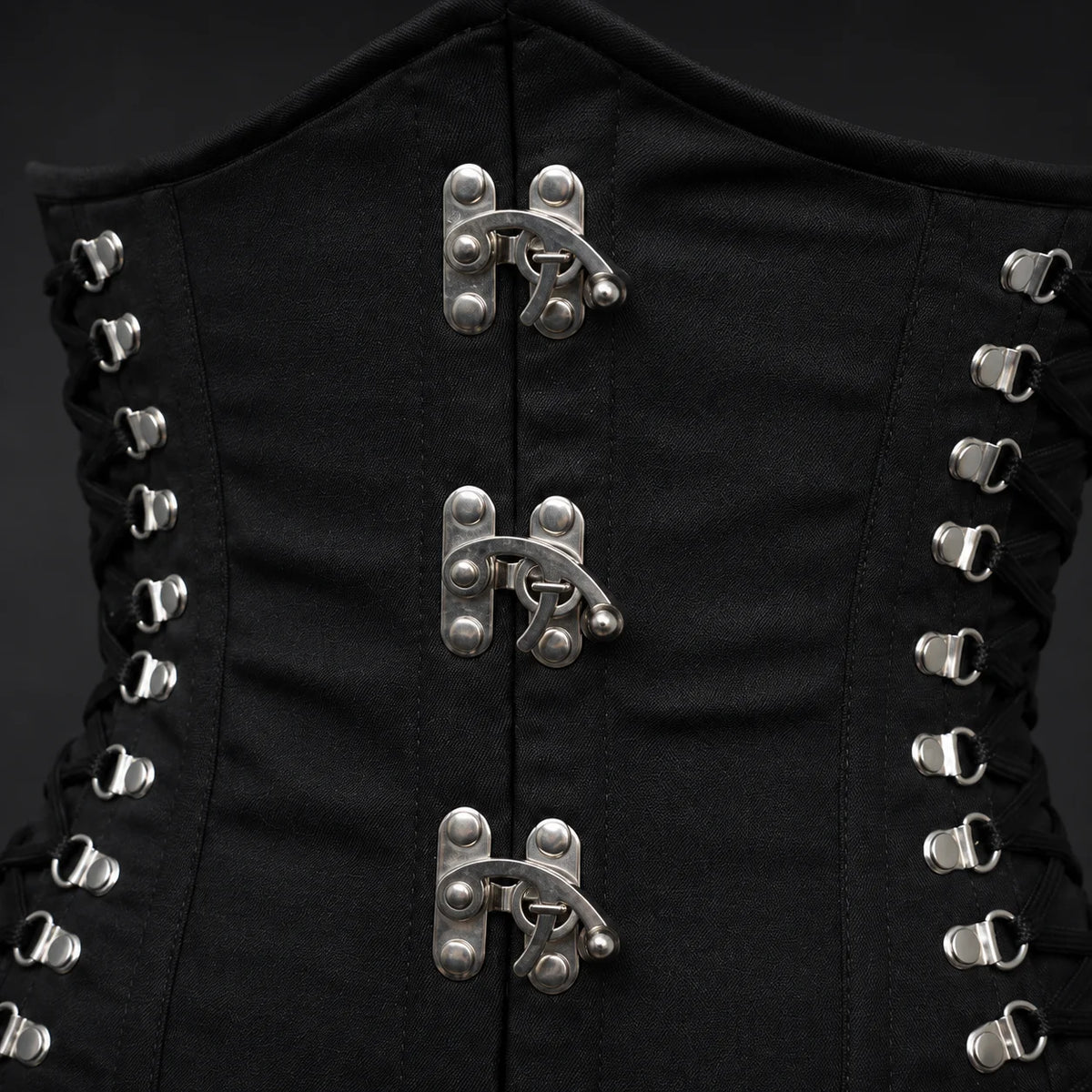 Close-up of black cotton corset front showing metal hook clasps, stitched panels, side lace fittings and matte fabric texture