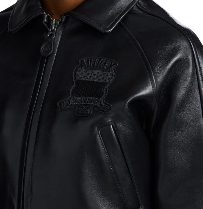 Close-up of women’s black leather bomber jacket showing structured collar, front zipper, chest emblem and welt pocket