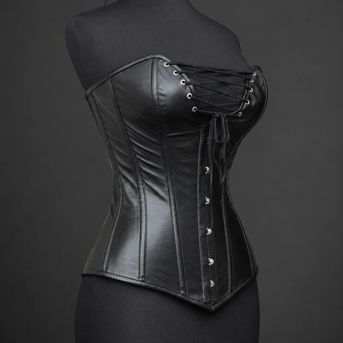 Three-quarter product angle showing smooth black leather, plunge bust lacing, curved panel seams and front busk detail.