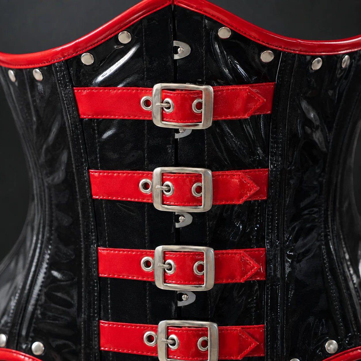 Close-up of glossy black PVC corset front showing red buckle straps, stitching, rivets and silver-tone hardware