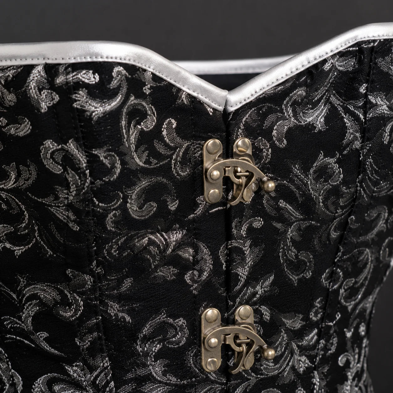 Close-up of black patterned corset front showing silver edging, detailed stitching and antique-brass-tone clasps