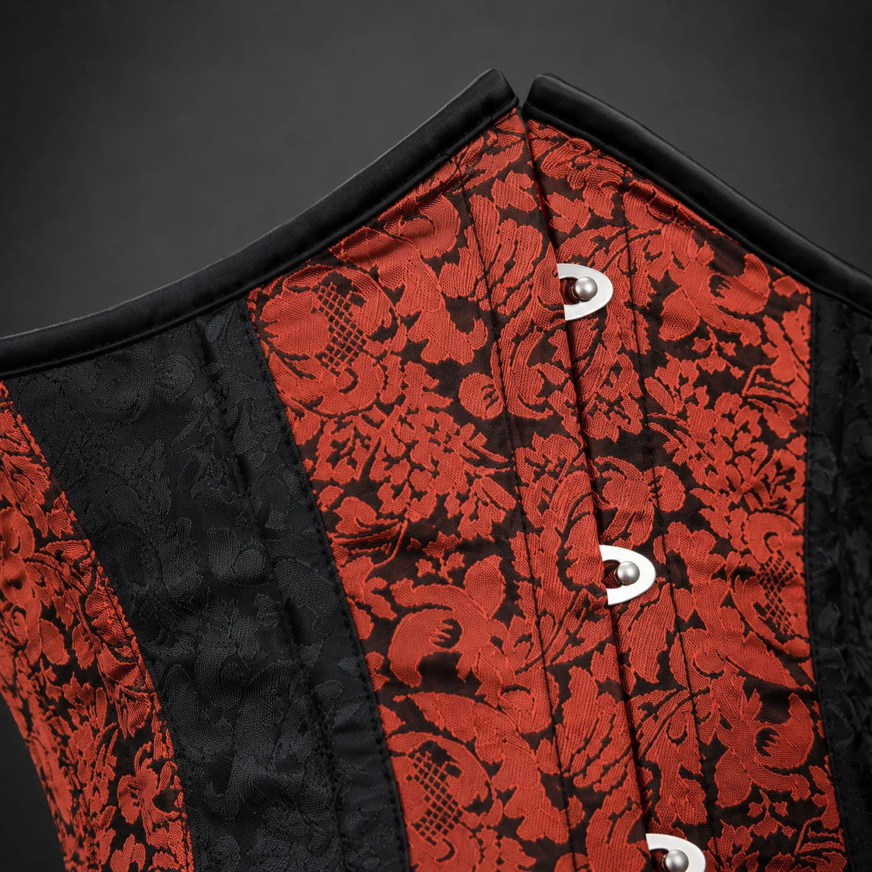 Close-up of red floral brocade corset fabric with black patterned panel, edge binding and silver-tone front busk hardware