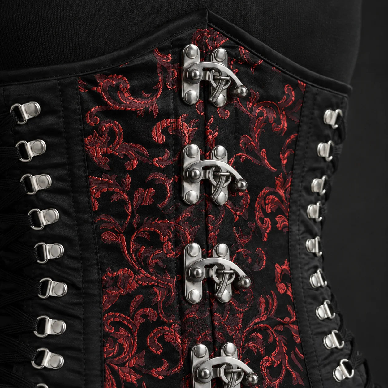 Close-up of red brocade corset front showing ornamental textile pattern, metal clasp hardware and side lacing