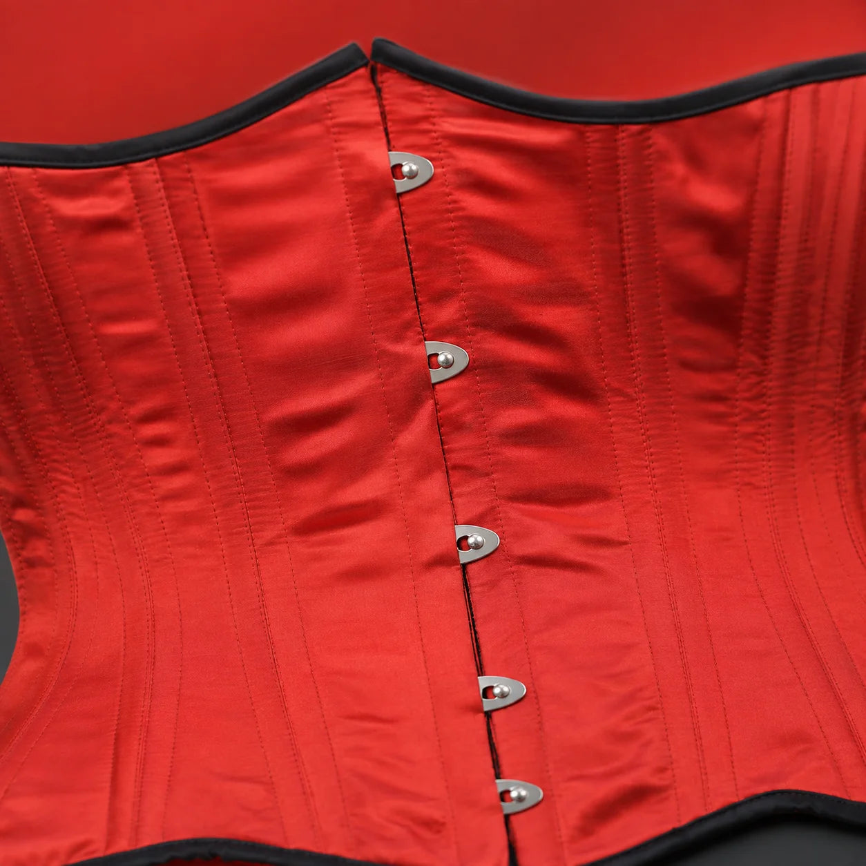 Close-up of glossy red satin corset showing detailed stitching, black edging and silver-tone front clasps